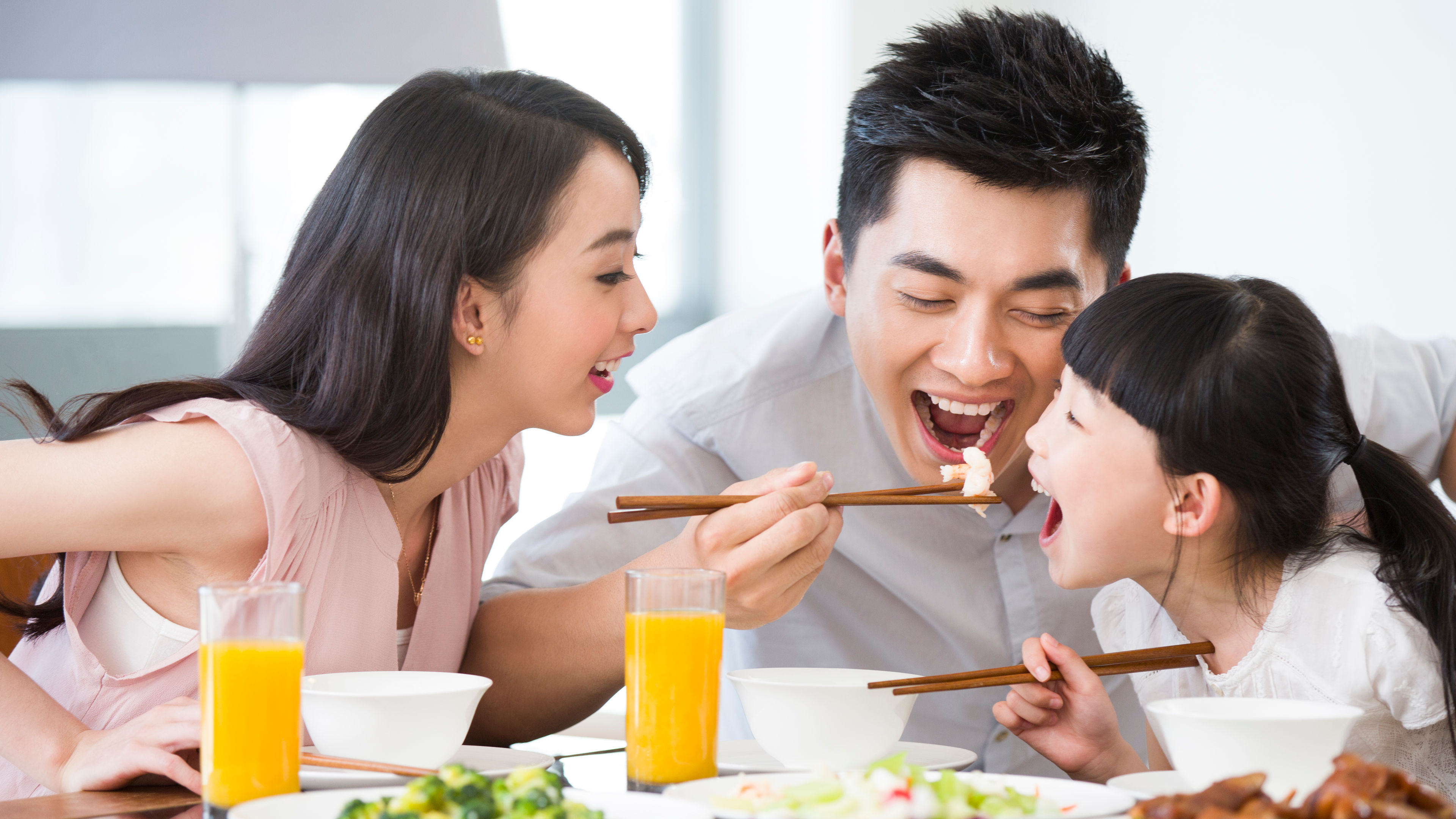 Asian family happily eating together, wearing light-colored clothes, with juice on the table.