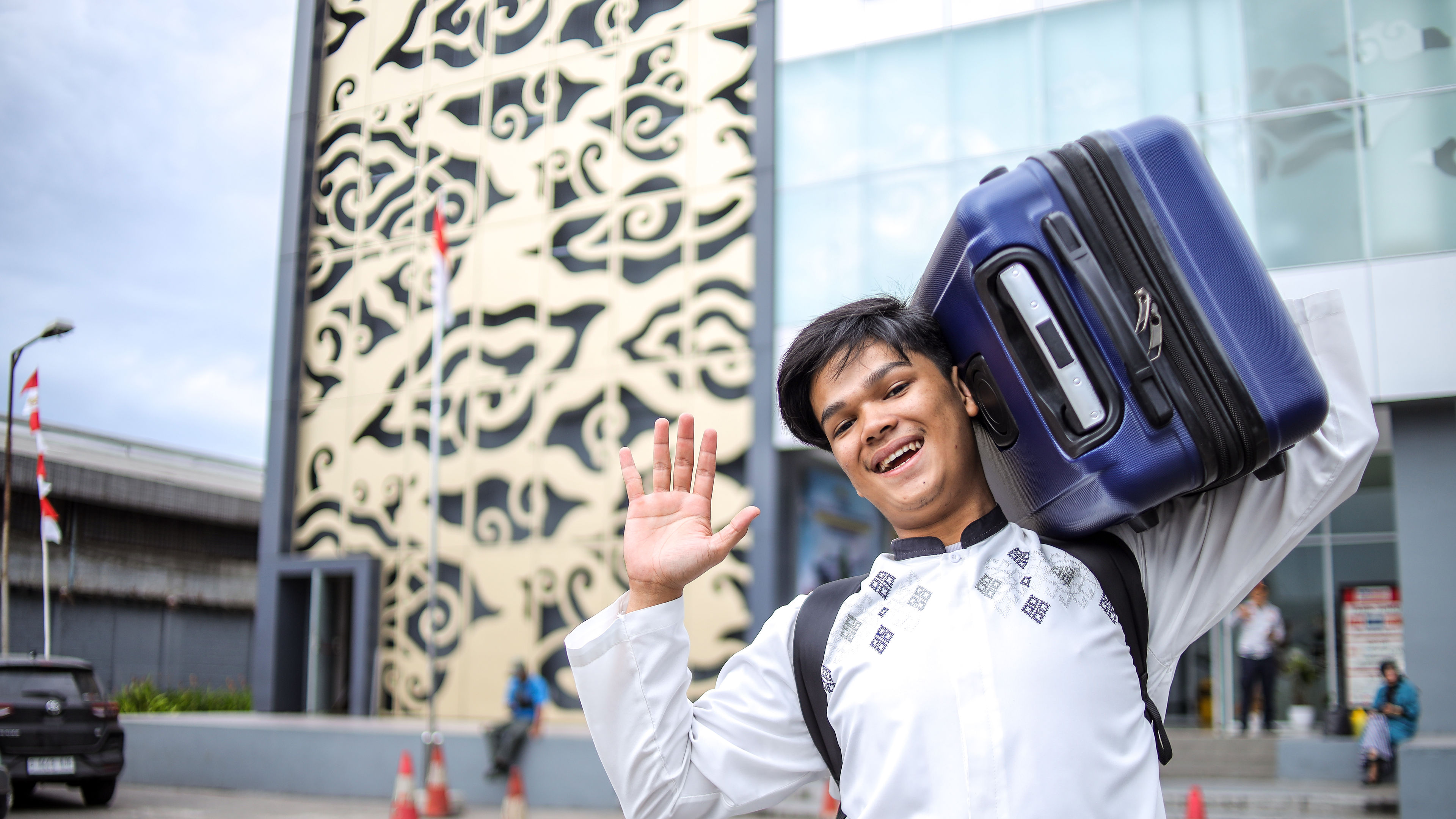 Young Asian Muslim Man Carrying Suitcase On Shoulder While Greeting By Waving Hand