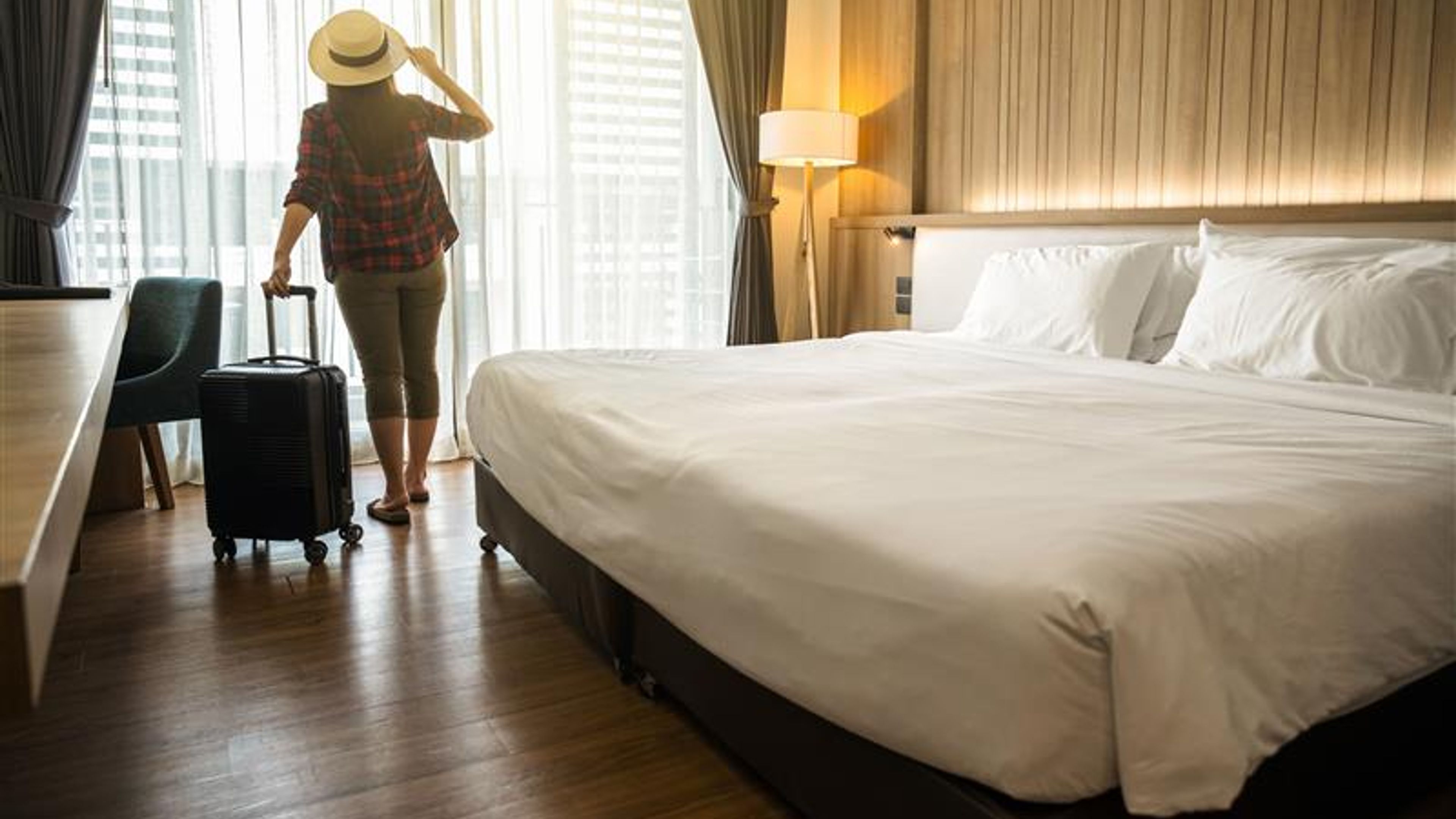 A woman stands by the window of her hotel room with her baggage beside her, smiling as she happily takes in the scenic view outside