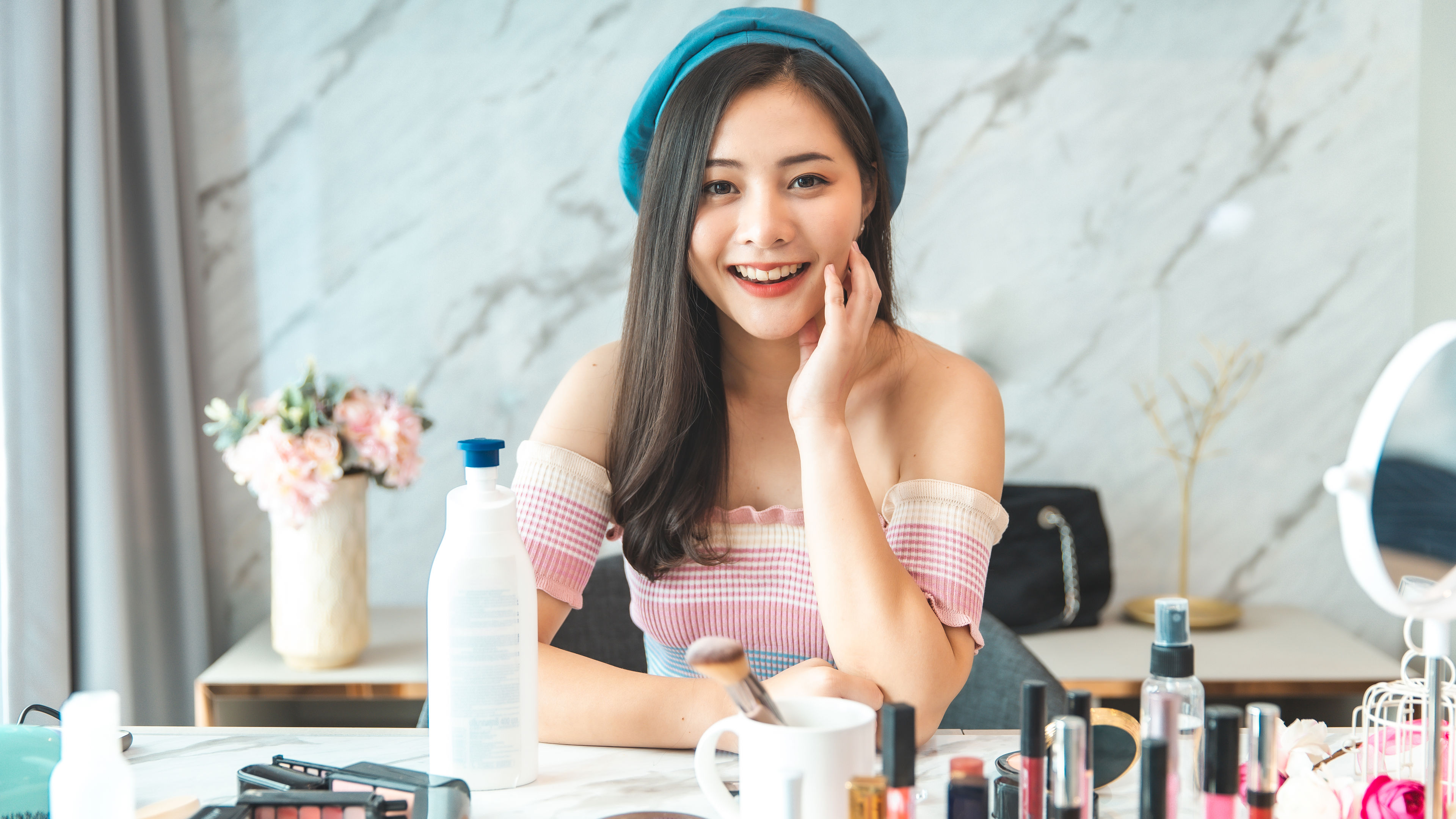 Woman smiling at a makeup table wearing a blue beret and pink off shoulder top with cosmetics and skincare products displayed.