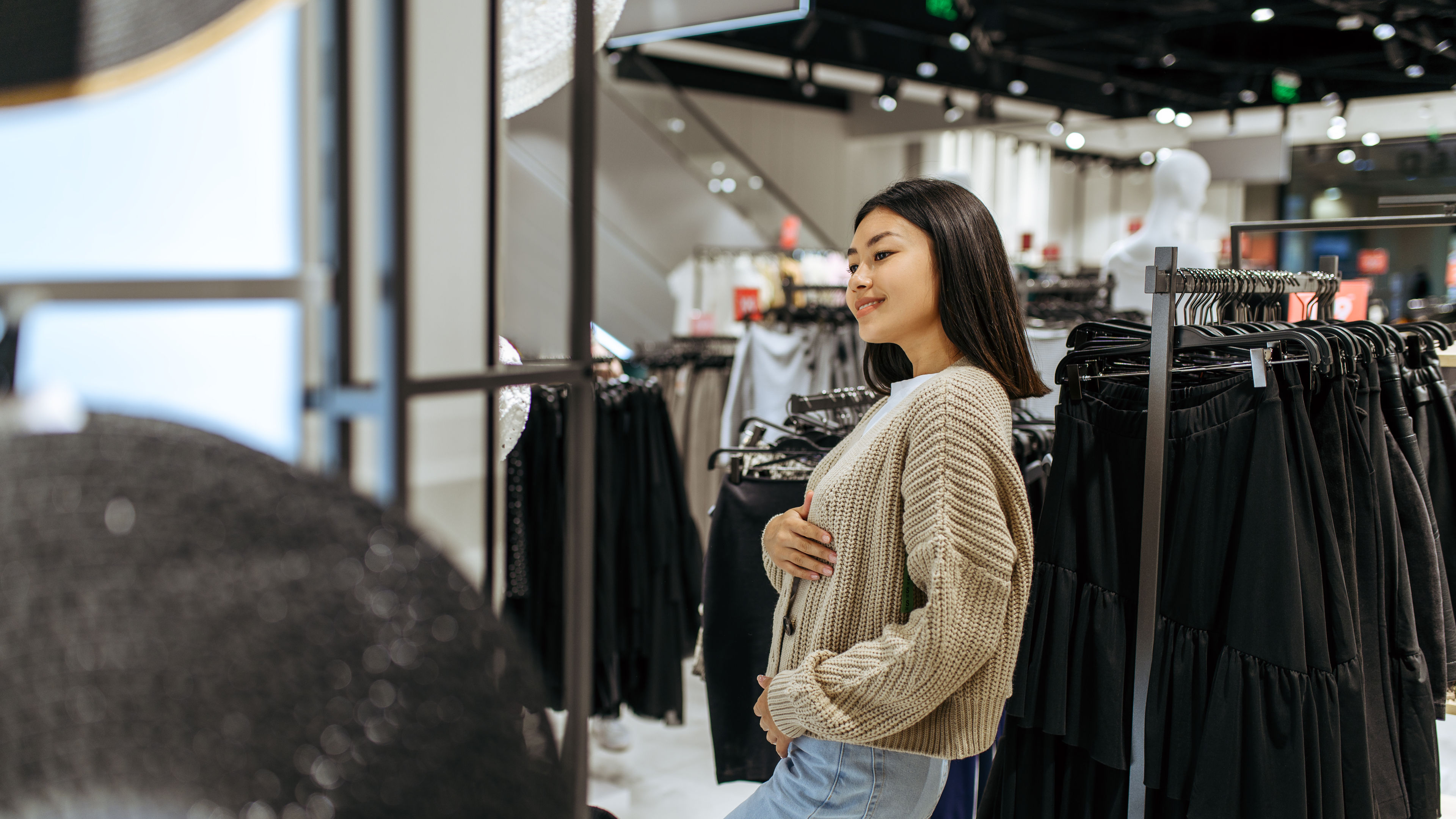 Young woman trying on a cardigan in a clothing store. Female people shopping in fashion boutiques, shopaholic, shoppers looking on garment 