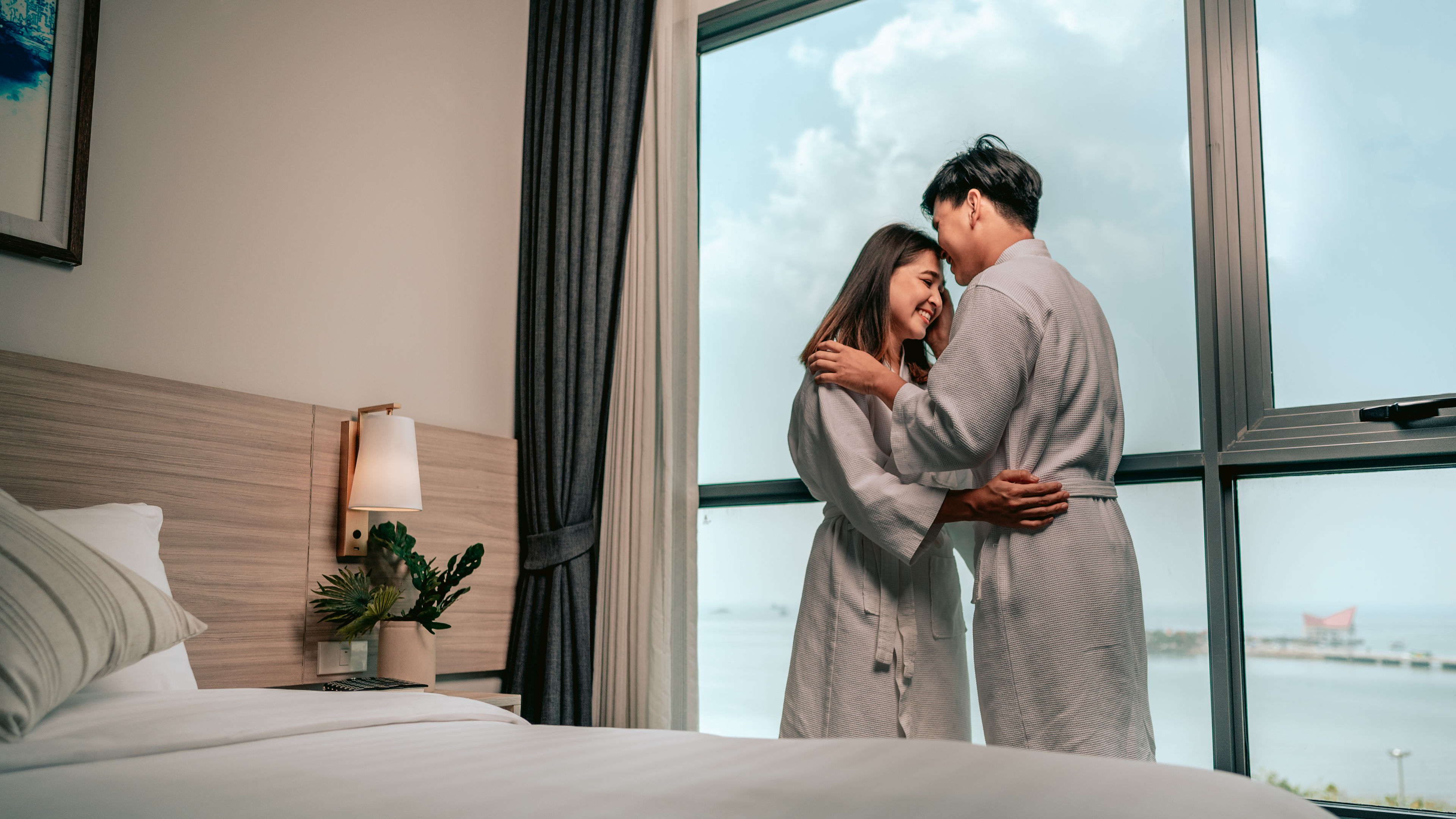 A couple in bathrobes sharing an intimate moment by a hotel room window with an ocean view.