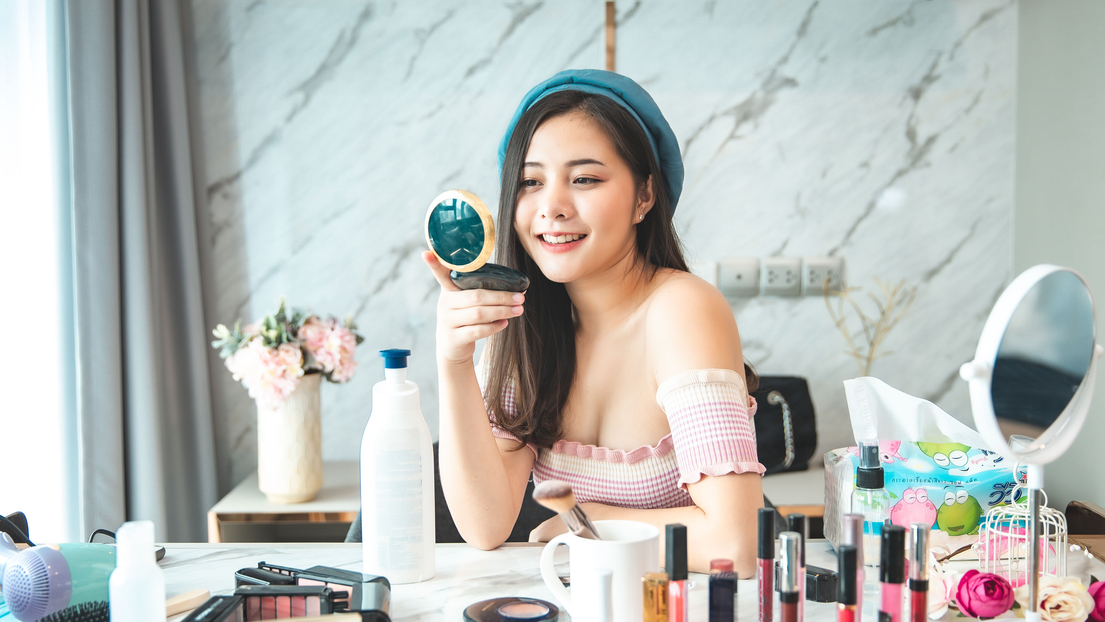 A portrait of a young Asian woman smiling as she reviews a cosmetics product, holding one product in her hand while others are placed on the table.