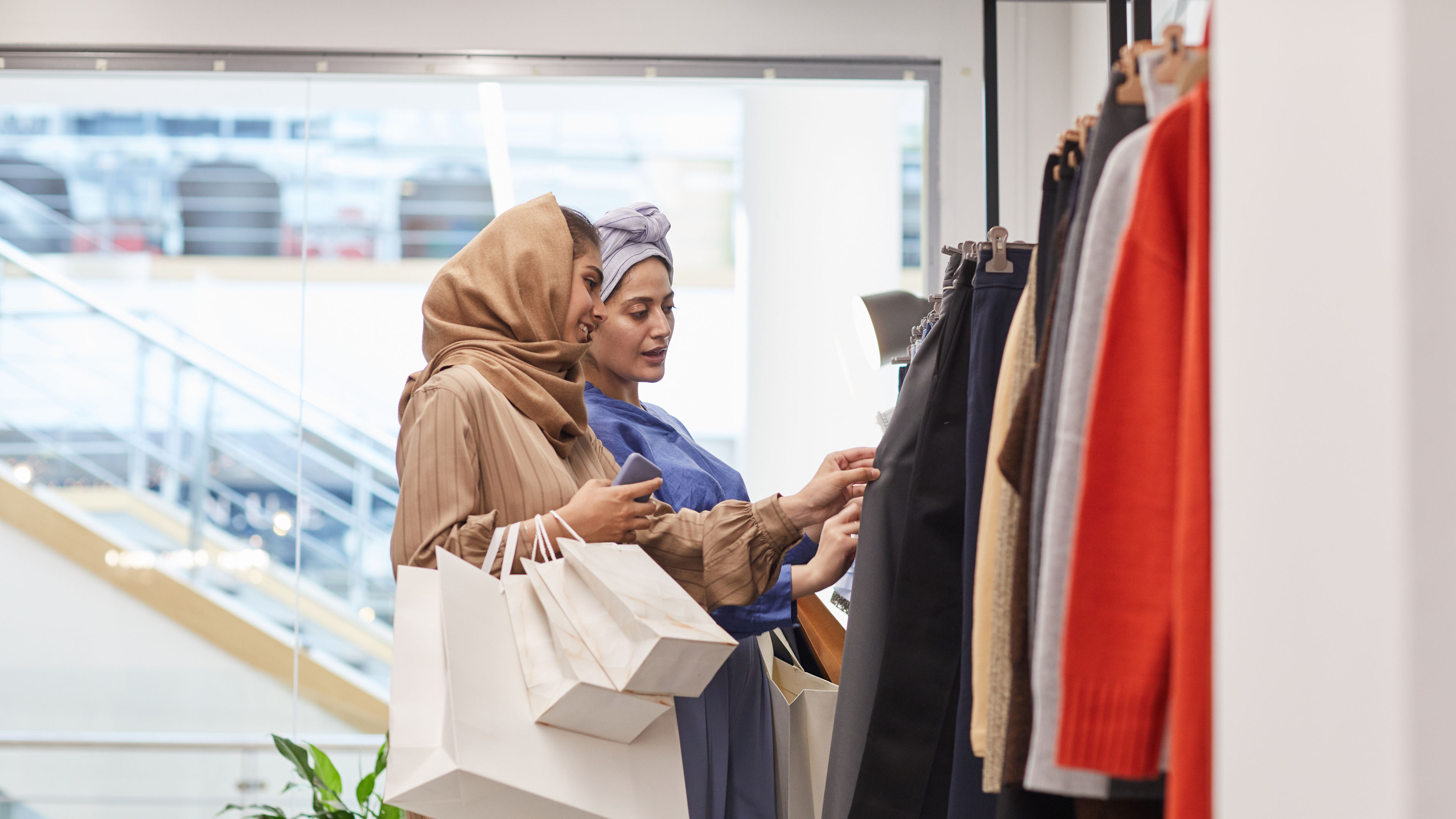 Two women shopping for clothes, examining garments on a rack while carrying multiple shopping bags.