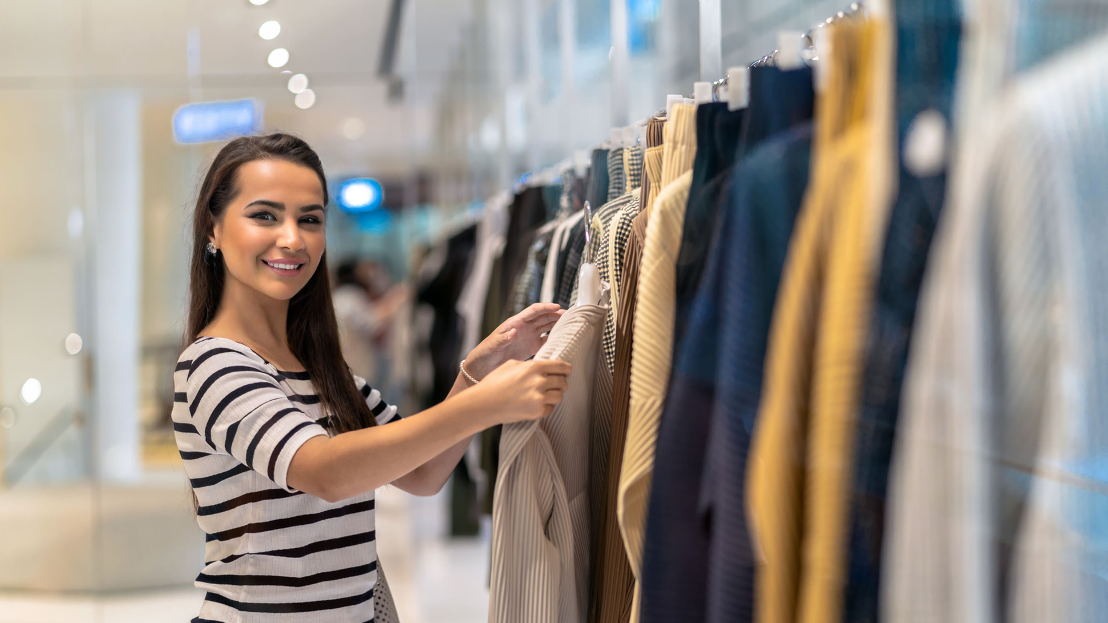 Woman shopping for clothes, smiling while browsing shirts on a rack in a store.