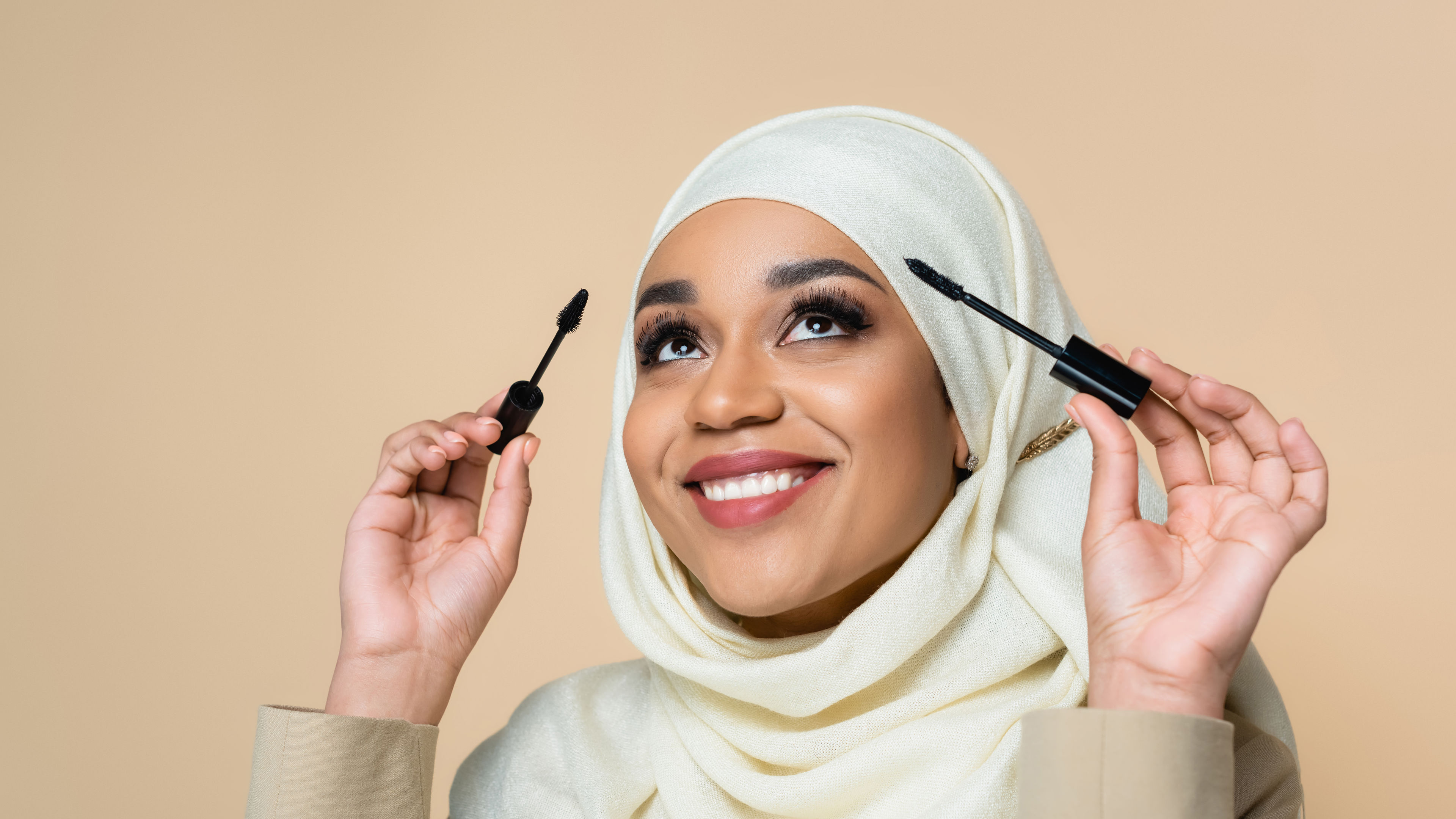 cheerful Muslim woman in hijab holding mascara brushes isolated on beige.