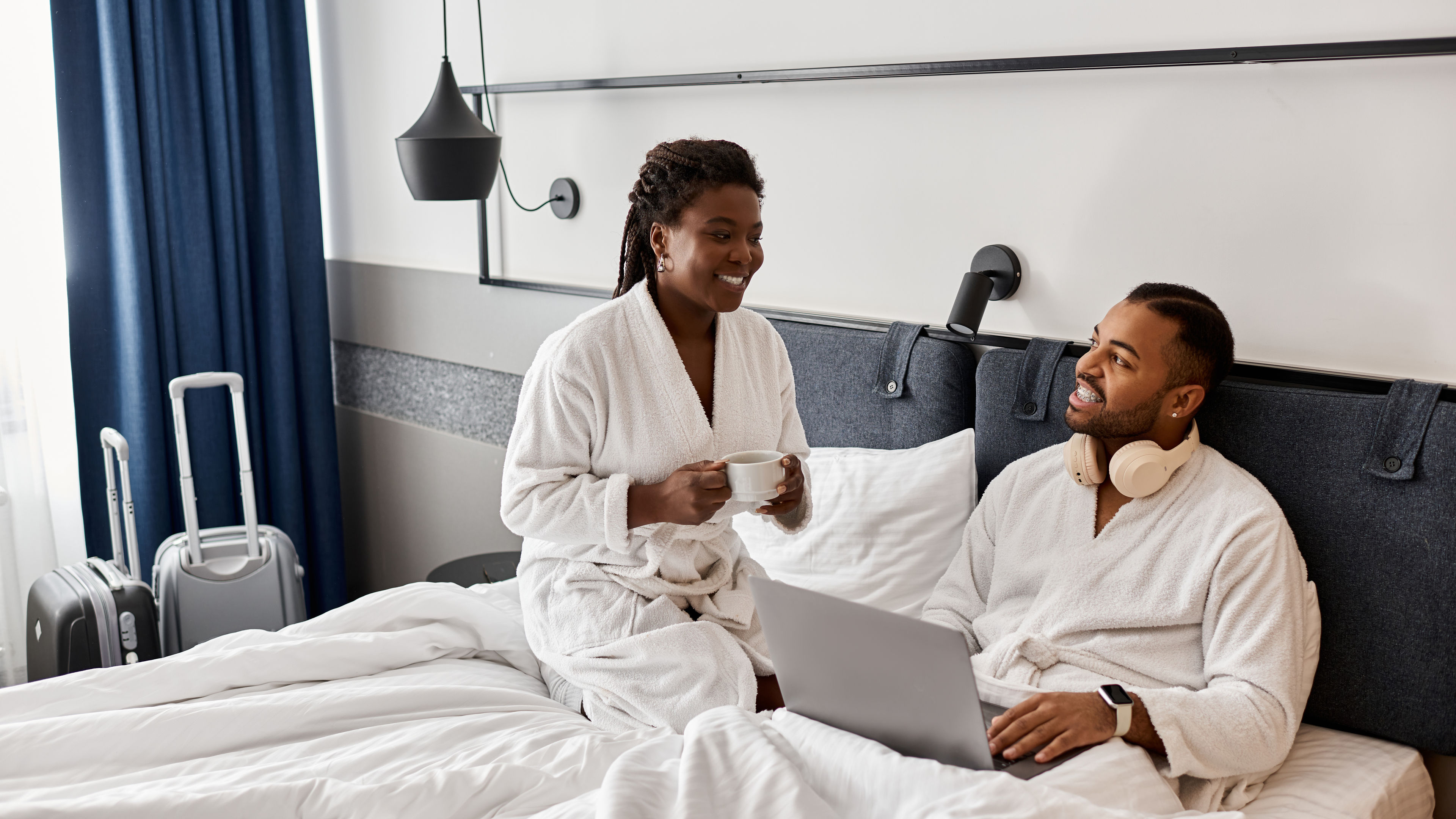 Smiling couple in bathrobes relaxing on a hotel bed, with luggage nearby and a cup of coffee in hand.