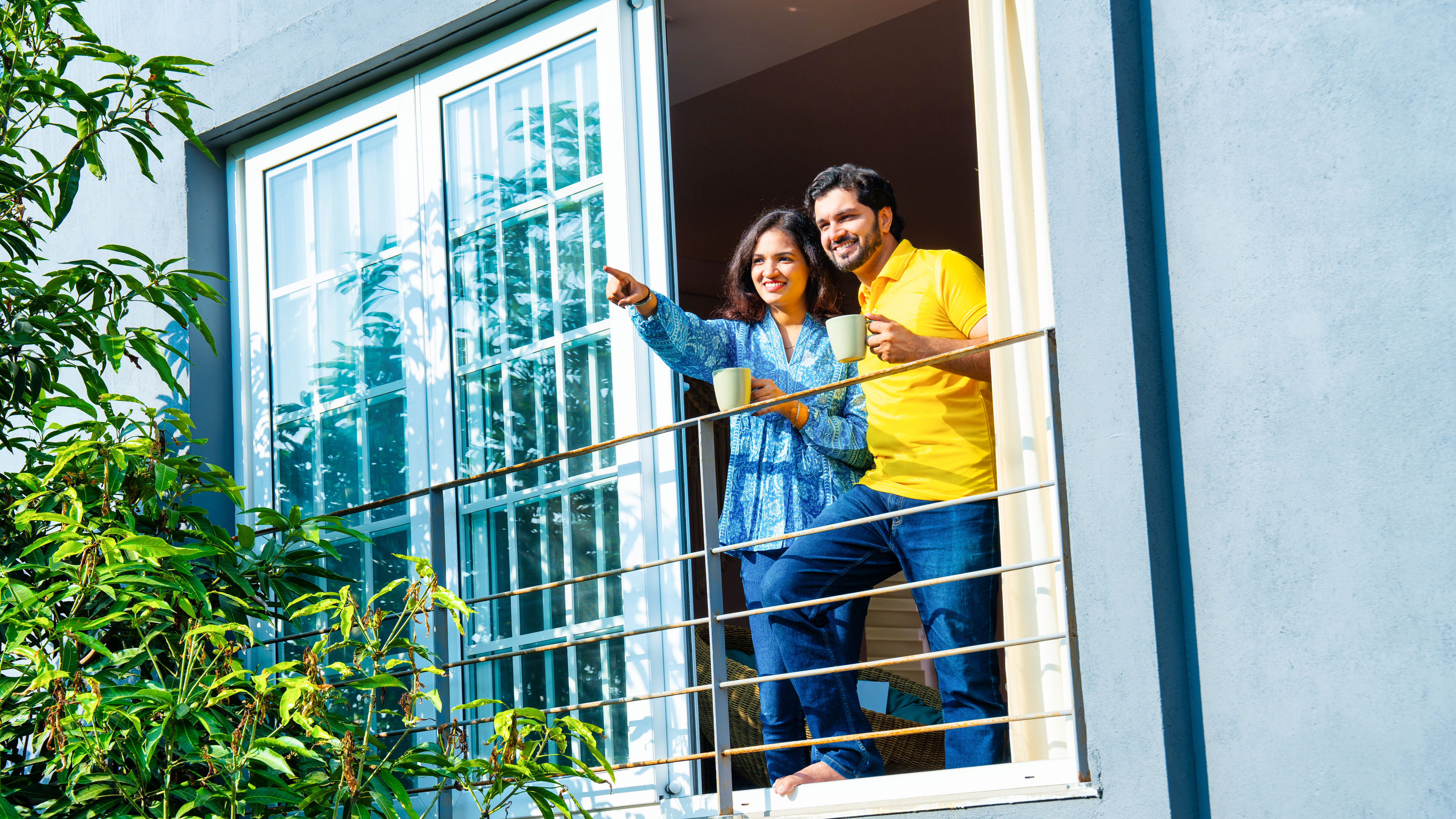A happy couple standing on a balcony, holding coffee mugs and enjoying the view together.
