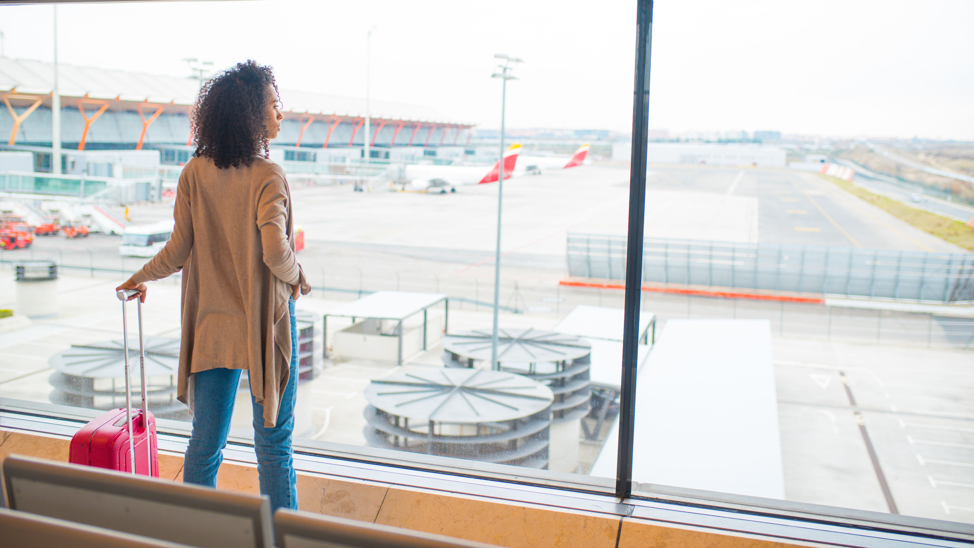 Black woman looking through airport window with a red suitcase.