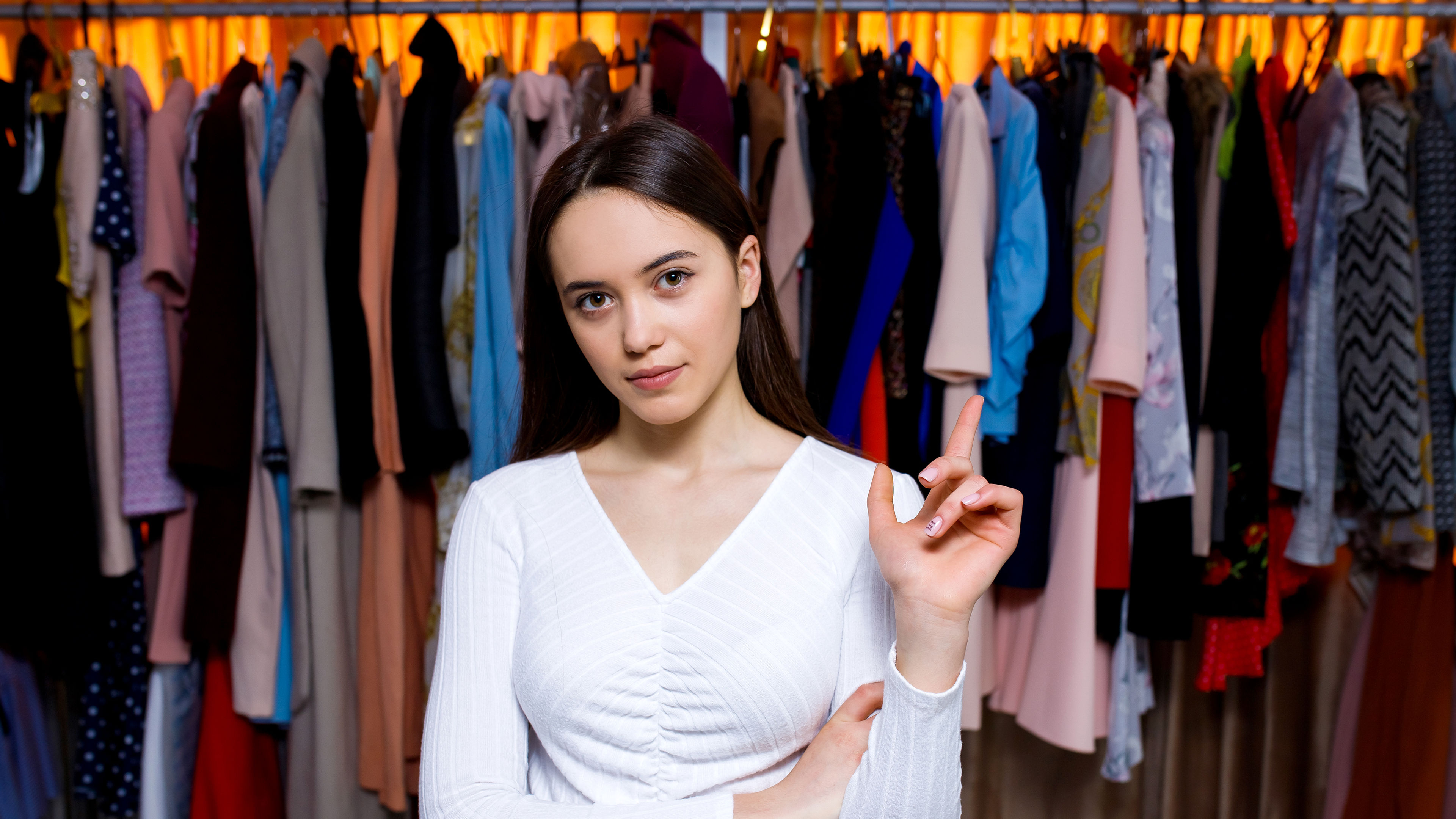 Young teenage girl stand store with clothes one hand on her chest other show things look in camera.