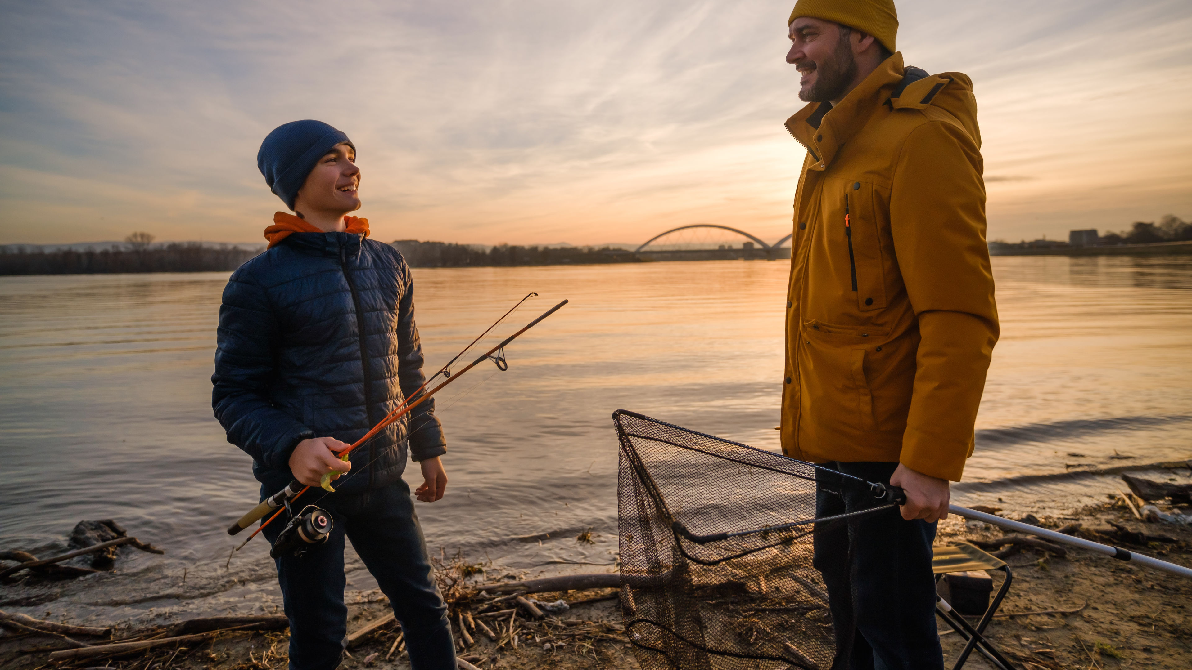 A father and son stand by a peaceful riverside at sunset.