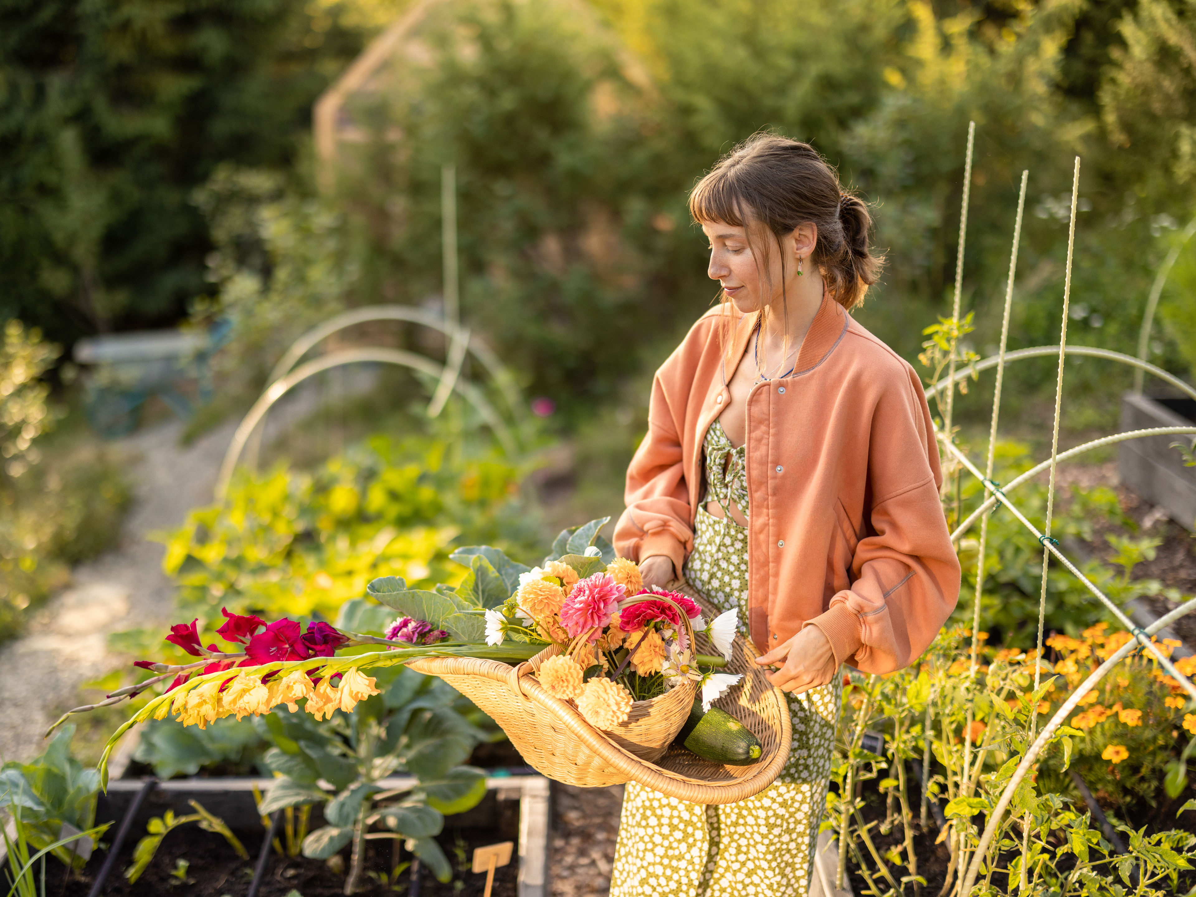 A young woman holding a basket of freshly picked flowers in a blooming garden at sunset.