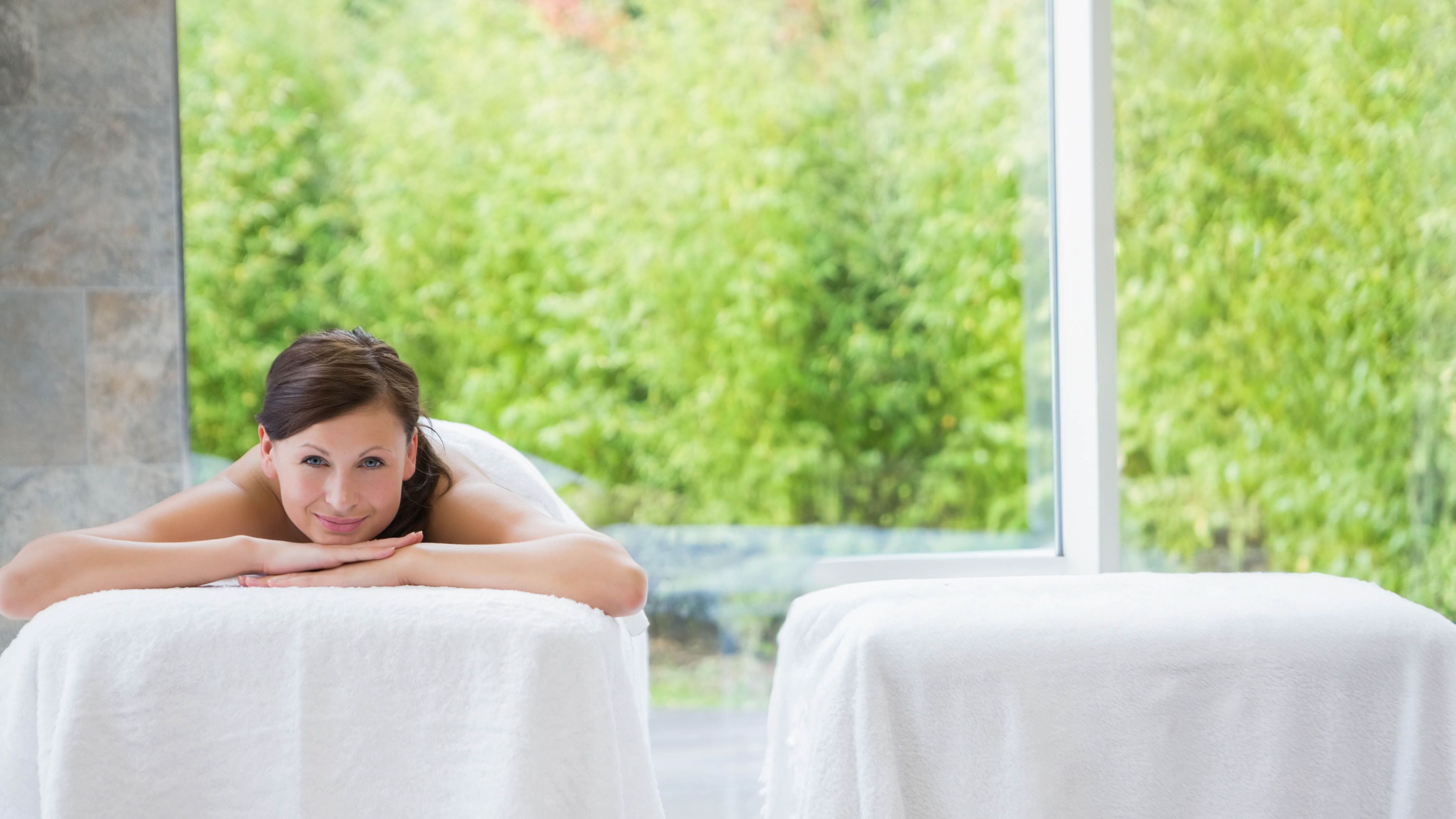 Female spa client lying prone on towel-covered massage table near glass wall with green shrubbery