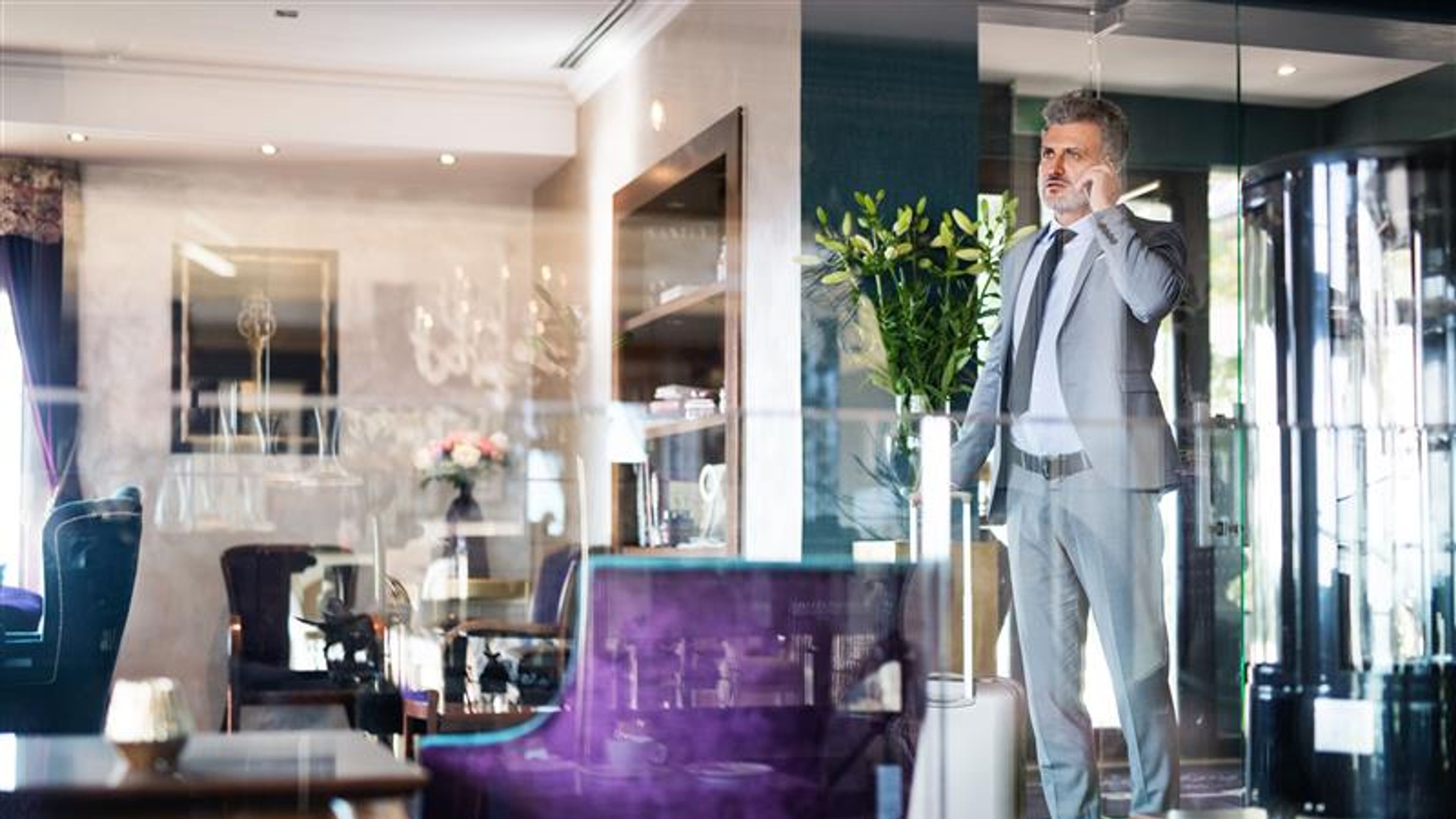 Handsome mature businessman with smartphone in a hotel lounge, making a phone call. Shot through glass