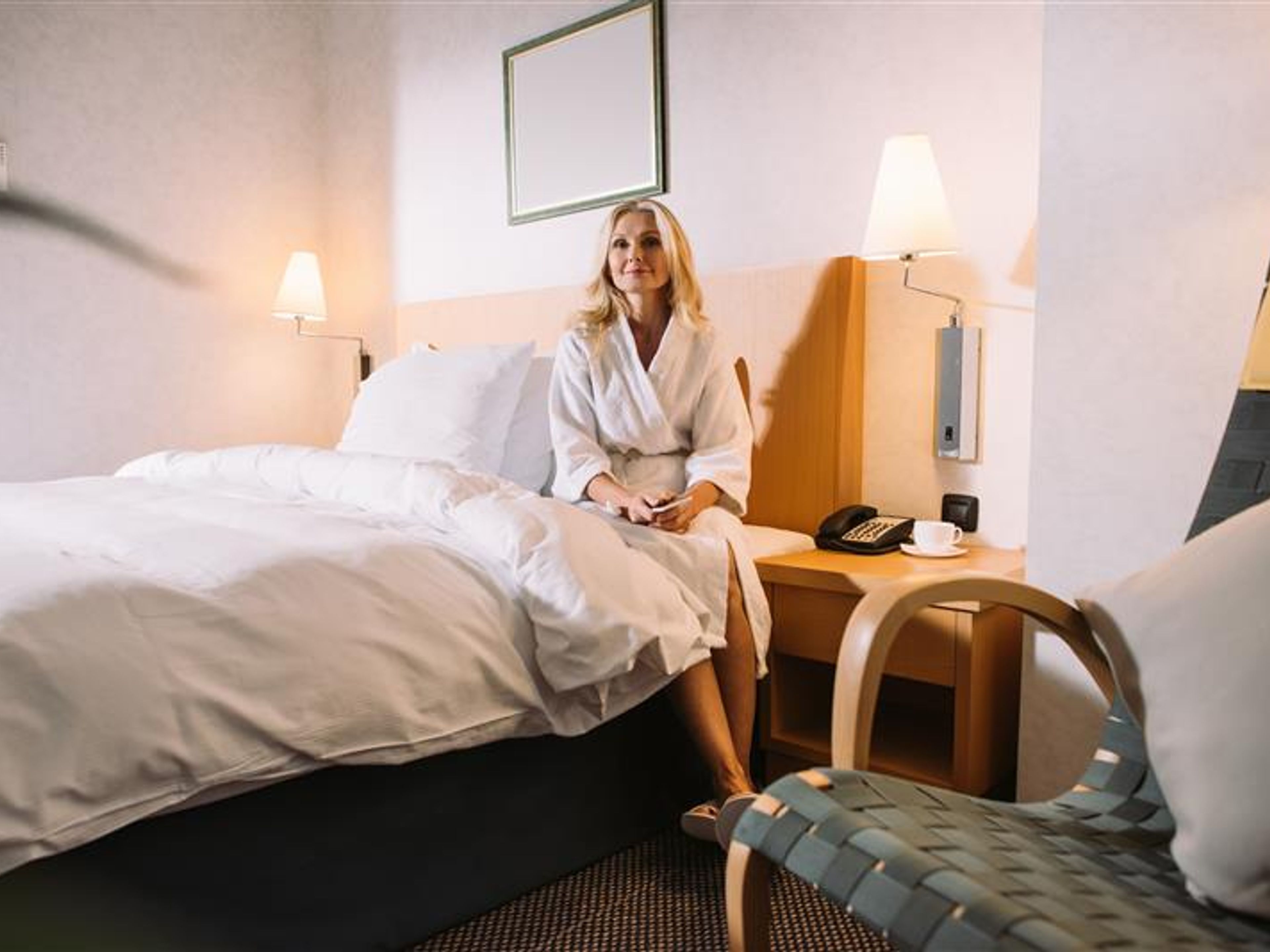 mature woman with smartphone in hands sitting on bed and looking away in hotel room